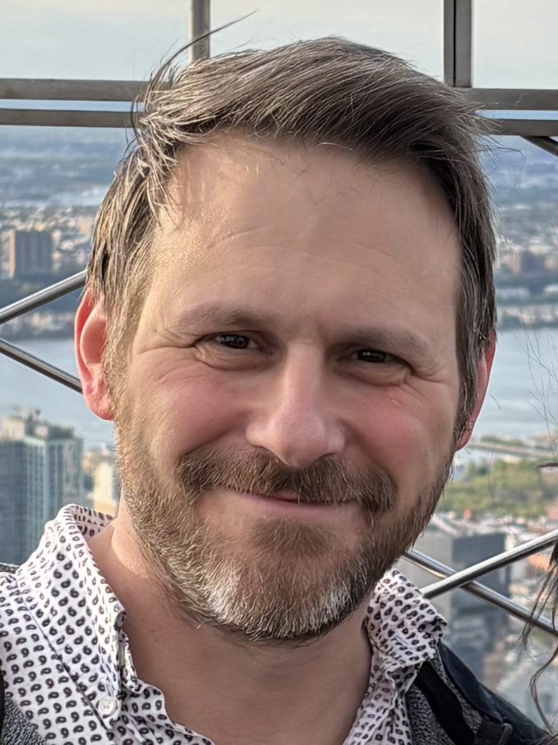 Brett Savoie smiling on a high observation deck with a city and water in the distance.